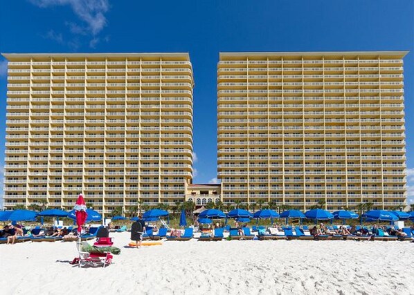 Deux grands bâtiments d’hôtel dotés de nombreux balcons, une plage de sable au premier plan et des parasols bleus.