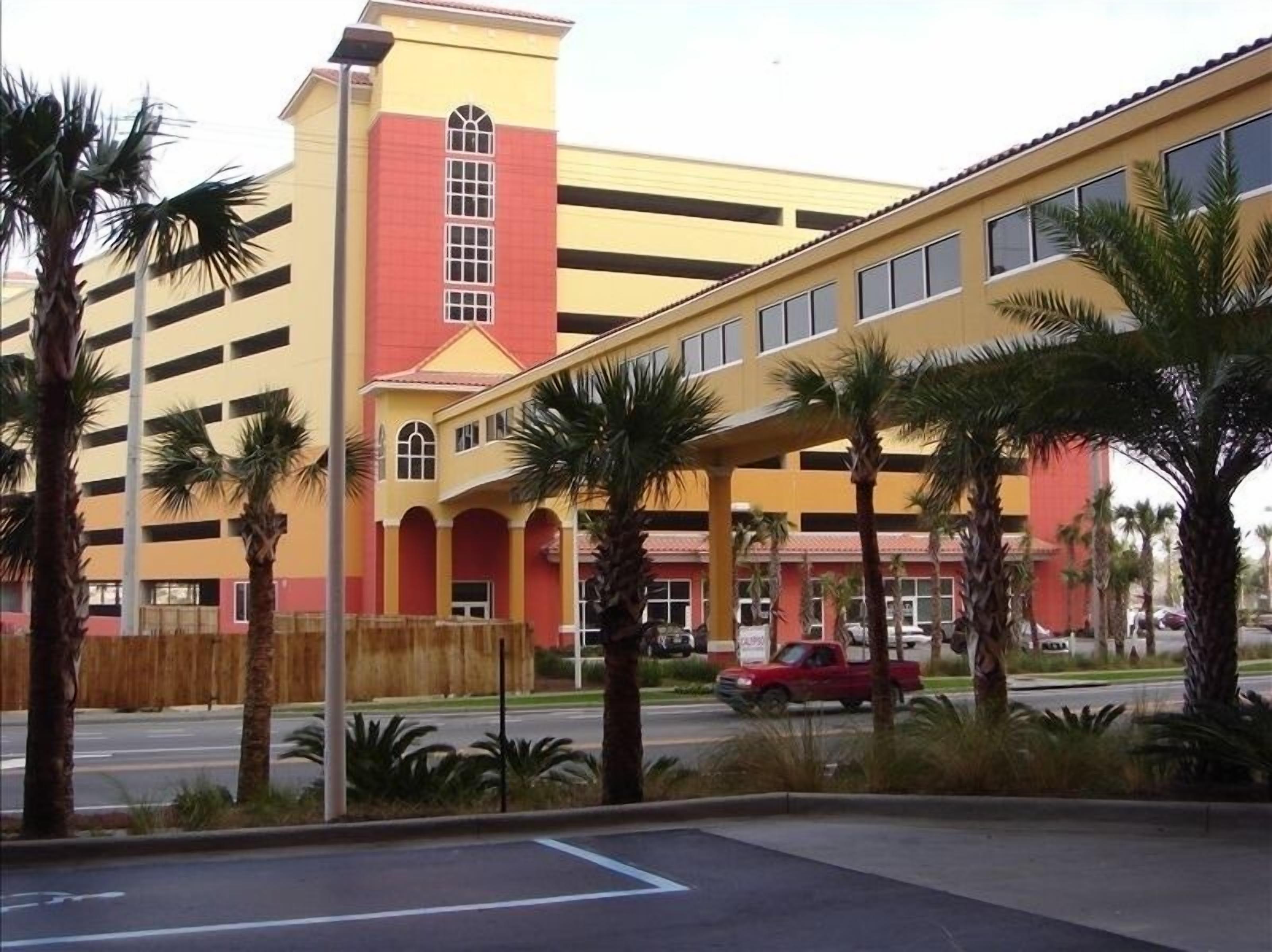 A multi-story parking garage with a yellow and red facade, palm trees, and a red vehicle on the street.