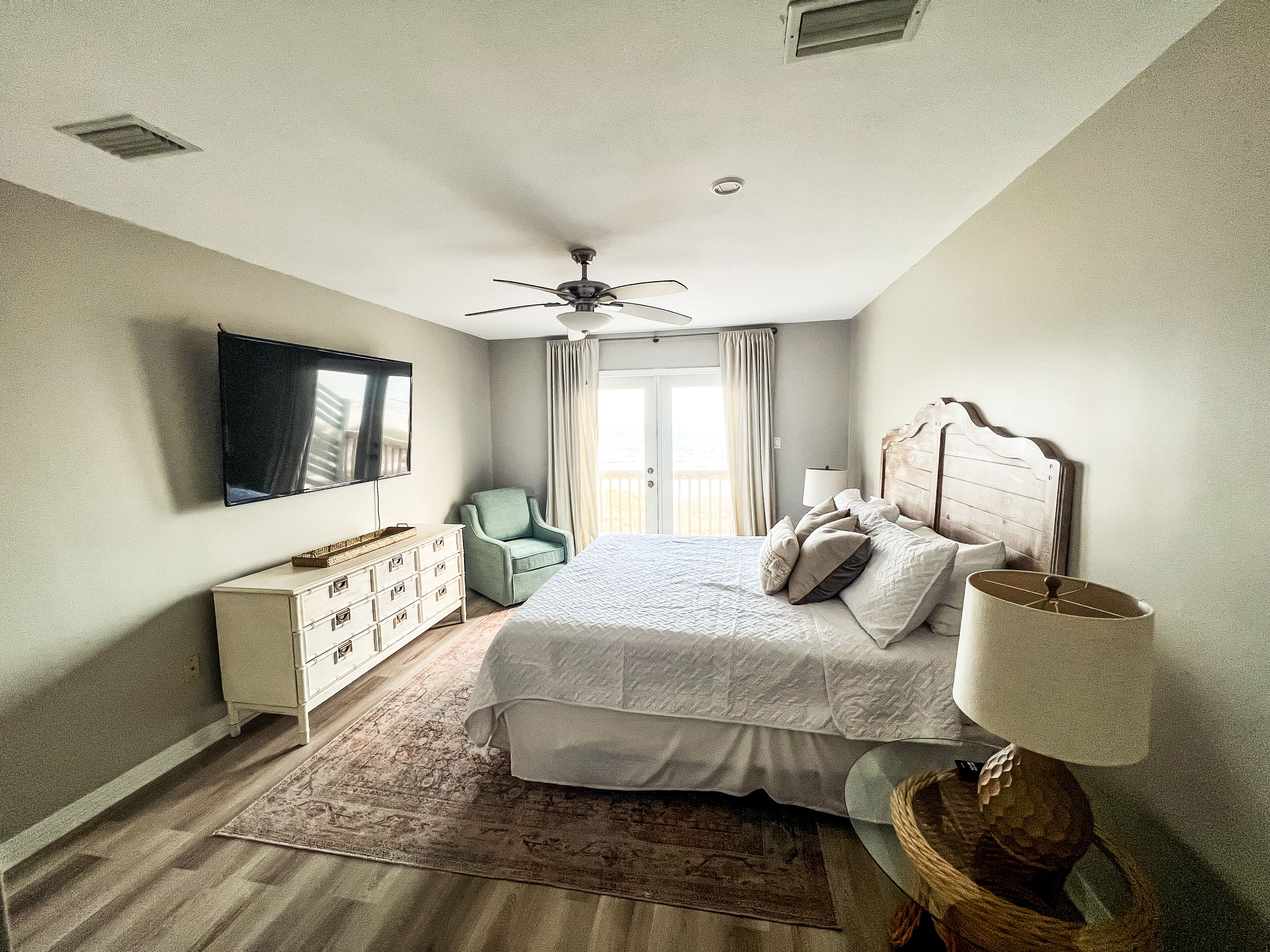 Une chambre à coucher comprenant un lit, une commode, une chaise, un téléviseur, un ventilateur de plafond et une fenêtre avec des rideaux.