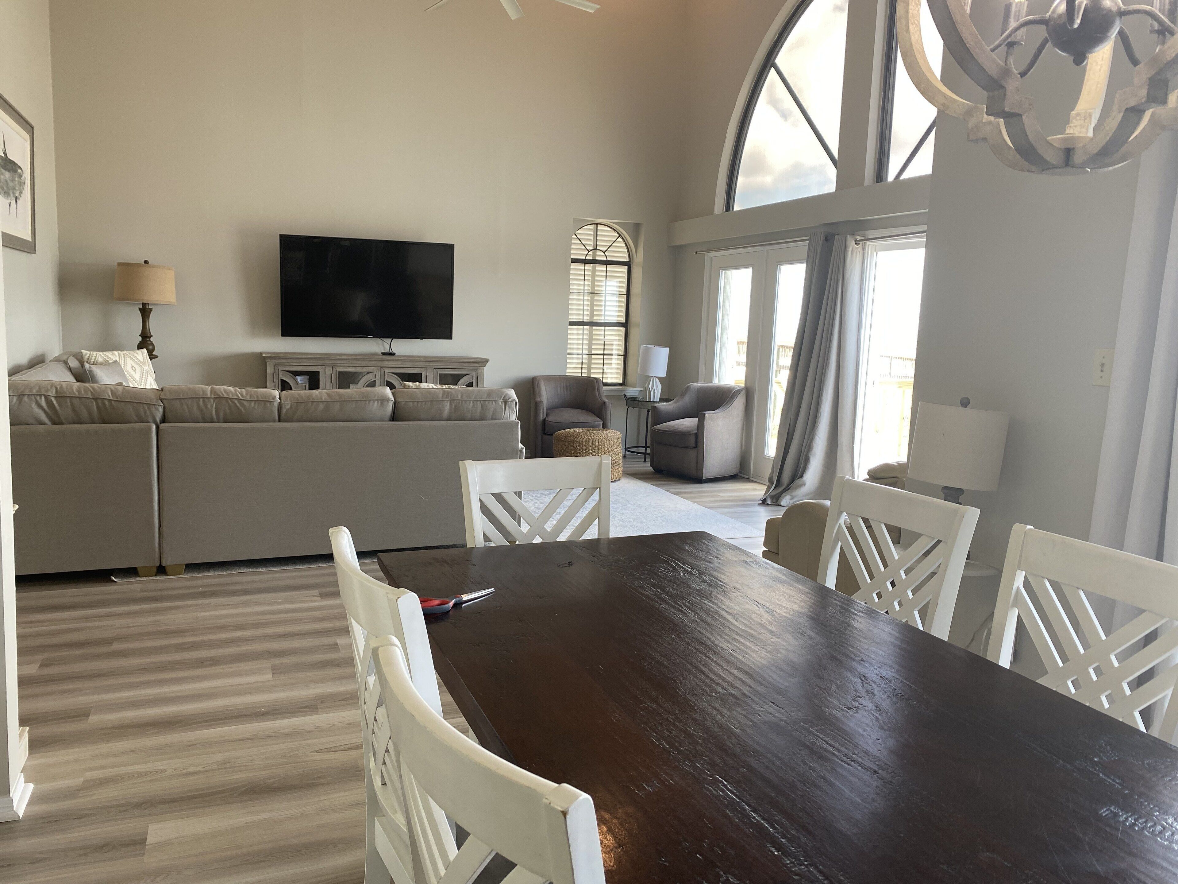 A spacious living room with a large dark wooden dining table, white chairs, a beige sofa, and a flat-screen TV mounted on the wall.