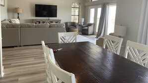 A spacious living room with a large dark wooden dining table, white chairs, a beige sofa, and a flat-screen TV mounted on the wall. - Relax With Walls Of Windows Directly On The Gulf! (Gulf Shores)