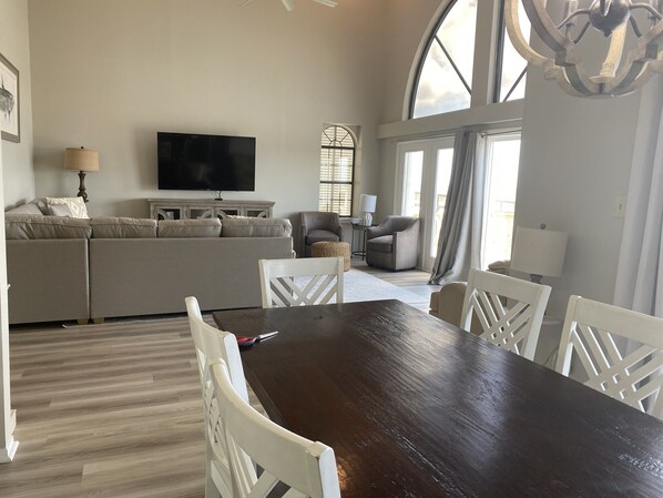 Uma sala de estar ampla com uma grande mesa de jantar de madeira escura, cadeiras brancas, um sofá bege e uma televisão de tela plana fixada na parede.