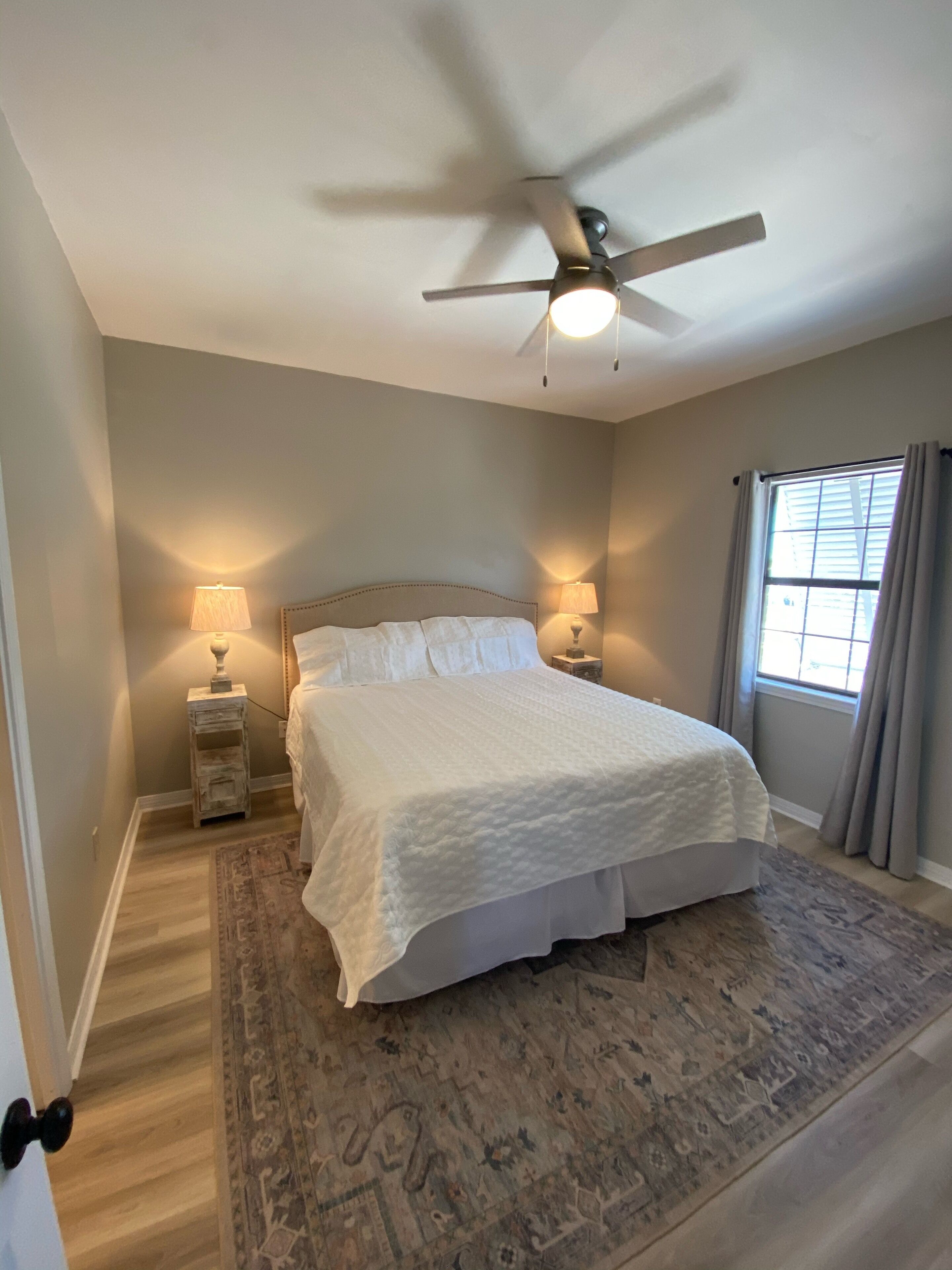 A bedroom with a bed, two bedside lamps, a ceiling fan, a window with curtains, and a patterned rug.