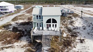 A beach house with a large deck, multiple chairs, and a staircase leading to a covered area. - Relax With Walls Of Windows Directly On The Gulf! (Gulf Shores)