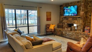 Una sala de estar con sofá, sillón reclinable, chimenea de piedra, televisor y vistas a árboles a través de amplios ventanales.