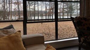 Una habitación con un ventanal que ofrece vistas a un área boscosa y una masa de agua. Hay un sofá con cojines y una silla con un cojín estampado.