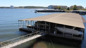 Ein Hafensteig am Wasser mit einer überdachten Bootsaufbewahrung, einer Fußgängerbrücke und Gebäuden im Hintergrund.