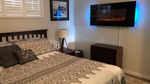 Ein ordentlich bezogenes Bett mit gemusterten Bettwäsche, eine Nachttischlampe, ein gerahmtes Bild an der Wand und ein Fernseher, der über einem Holzschrank angebracht ist.