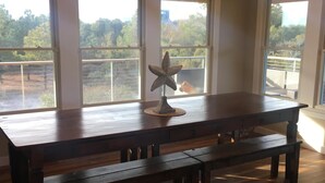 A spacious room with a large wooden dining table, a bench, and a decorative starfish sculpture. Three large windows provide views of a balcony and greenery outside.