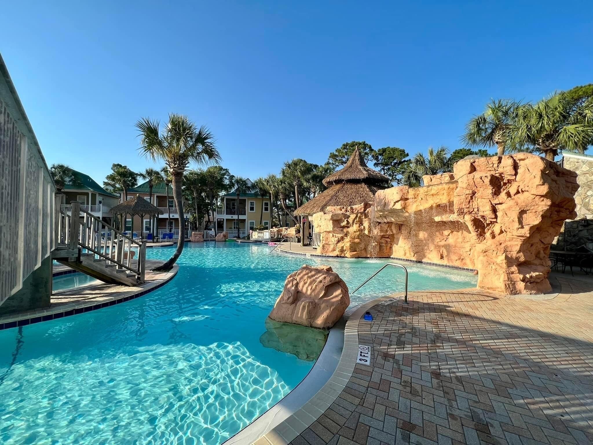 A swimming pool with clear blue water, surrounded by a brick deck and palm trees.