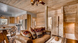 A cozy living room with a wooden ceiling, a sofa with patterned cushions, a coffee table, and a dining area with a wooden table and chairs.