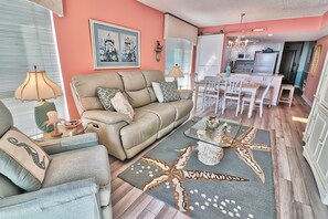 A living room with a beige sofa, a glass coffee table, and a star-patterned rug. There is a dining area with white chairs and a table.