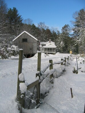 A wooden cabin and a house covered in snow, surrounded by trees.