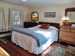 A bedroom with a large bed, a dresser, a nightstand, a lamp, a mirror, and a window with curtains.