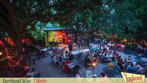 Un área de comedor al aire libre con mesas y sillas, un escenario con artistas y luces colgantes.