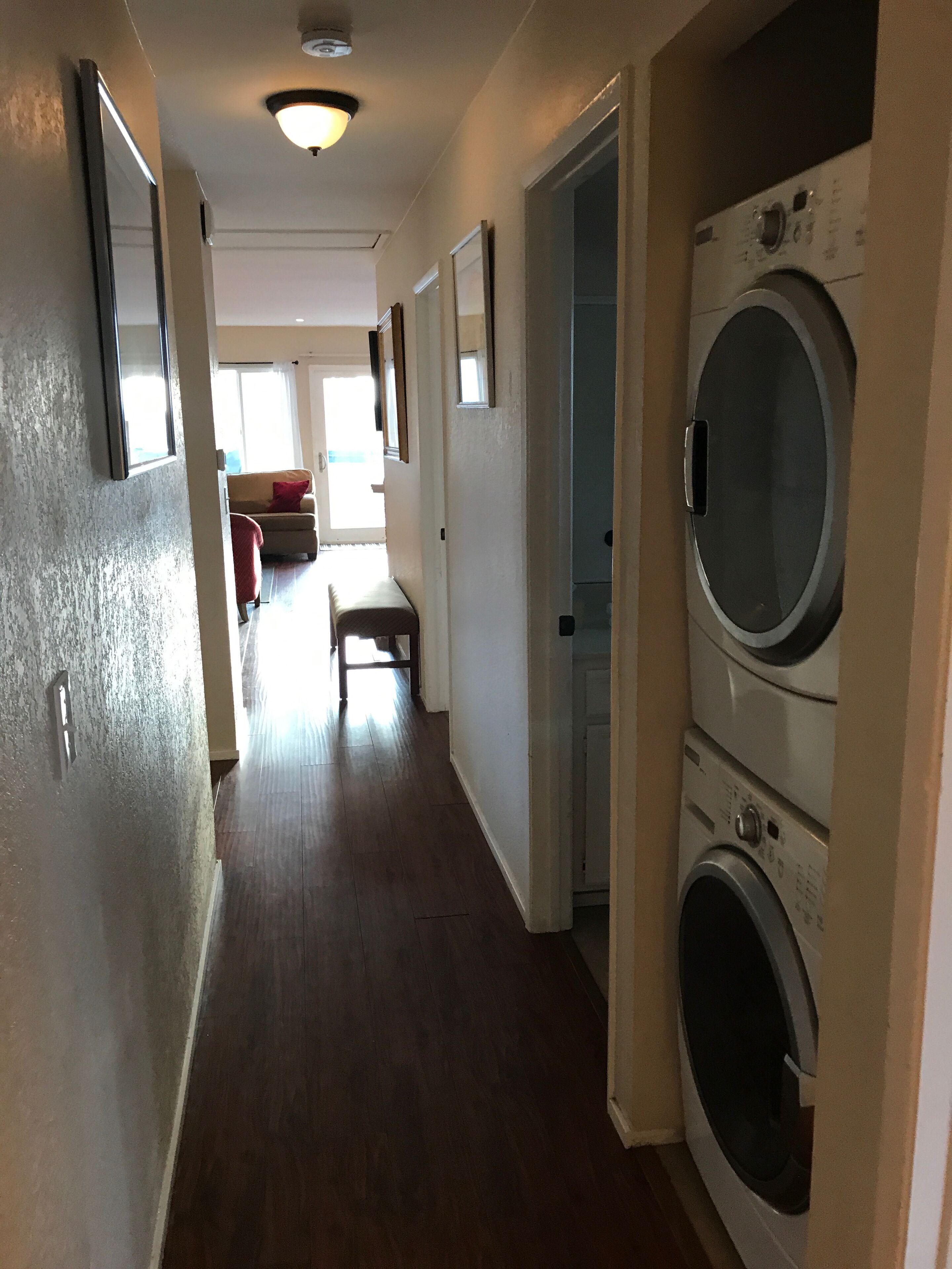 Una sala de bugaderia amb una rentadora i assecadora de càrrega frontal, un passadís amb terres de fusta, i una porta que condueix a una zona d'estar amb un sofà i una finestra.