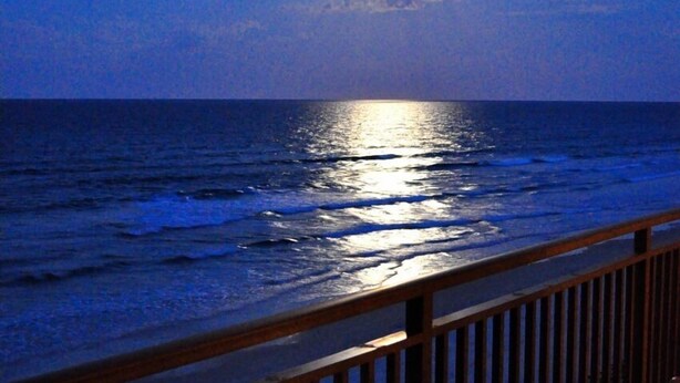 Ein Balkon mit Tisch für zwei Personen, mit Blick auf das Meer bei Nacht, mit sichtbarem Mond und Sternen.