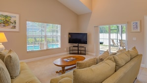 A spacious living room with a large sofa, a coffee table, and a TV. There are large windows with blinds, a ceiling fan, and a view of a pool and deck area. - Olde Florida Style, 4 Bedrooms 3 Bath Pool\Hot Tub - Free Pool and Spa Heat! (Naples)