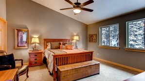 Une chambre à coucher comprenant un lit, des tables de chevet, un bureau, une chaise, un banc, un ventilateur de plafond et de grandes fenêtres donnant sur un paysage enneigé.