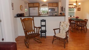 Une cuisine avec des meubles de rangement en bois, un four à micro-ondes et un réfrigérateur. On y trouve deux fauteuils à bascule et une table à manger avec des chaises.