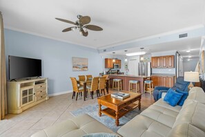 Sala de estar com televisão, sofá, mesa de centro e área de jantar com cadeiras, além de cozinha ao fundo.