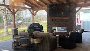 Une terrasse extérieure couverte, avec des poutres en bois, des meubles en osier, une cheminée en pierre et un barbecue.