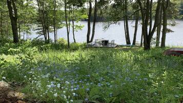 A lakeside setting with a table and chairs set up for outdoor dining, surrounded by trees and wildflowers.