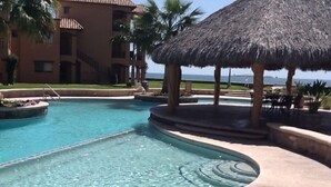 Una piscina con techo de paja, palmeras y un edificio al fondo.