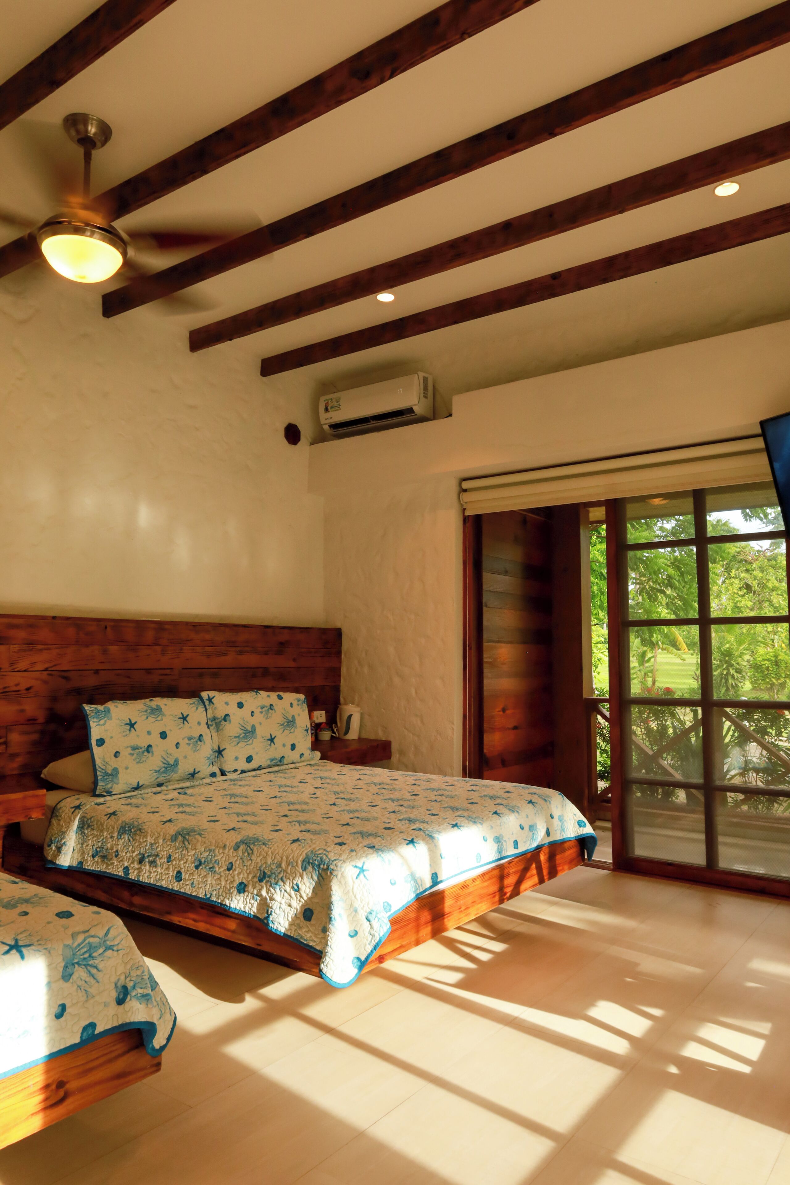 Quarto com cabeceira de madeira, cama com colcha estampada, ventilador de teto e janela com vista para o verde.
