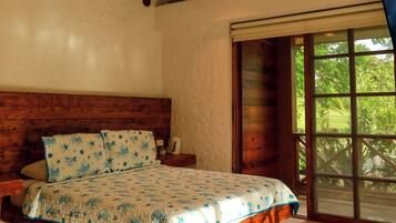 Quarto com cabeceira de madeira, cama com colcha estampada, ventilador de teto e janela com vista para o verde.
