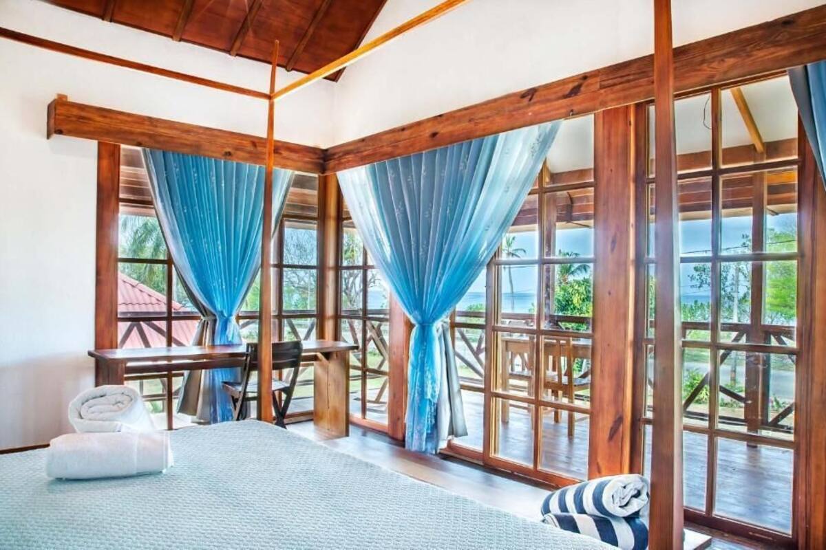 Una habitación con cama, una mesa y sillas, ventanas amplias con cortinas y vistas al exterior con vegetación.