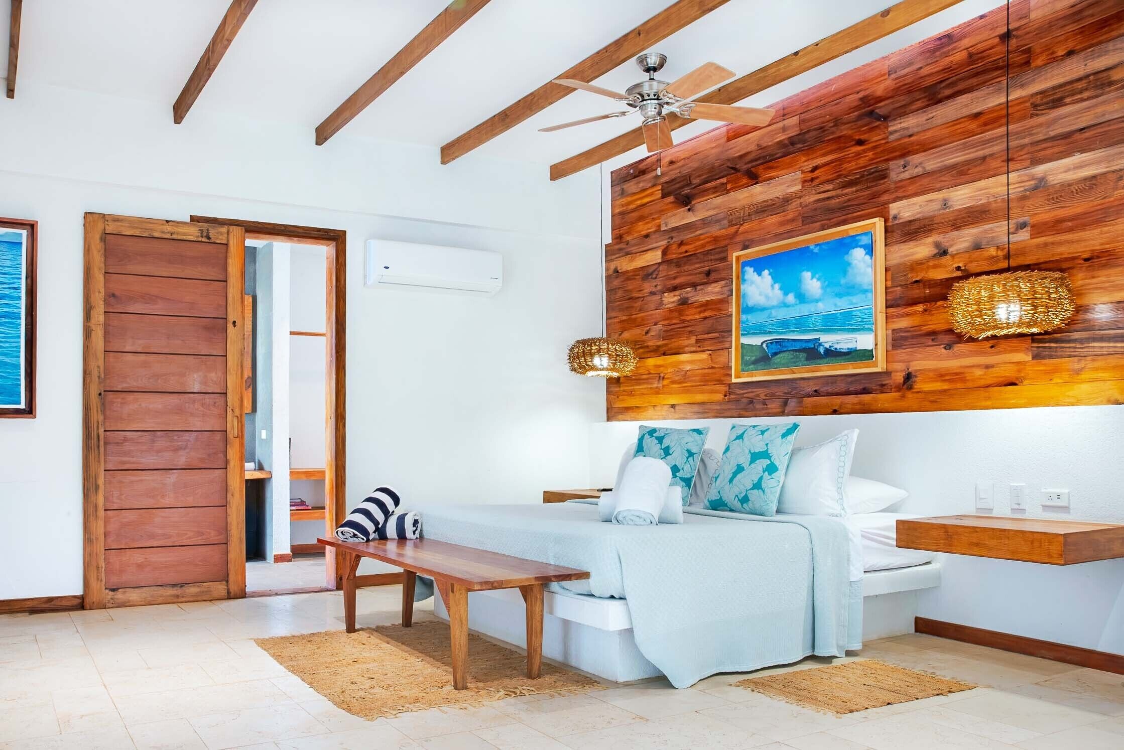 Un dormitorio con techo de madera, una cama con colchón blanco, un banco de madera y una estantería de pared.