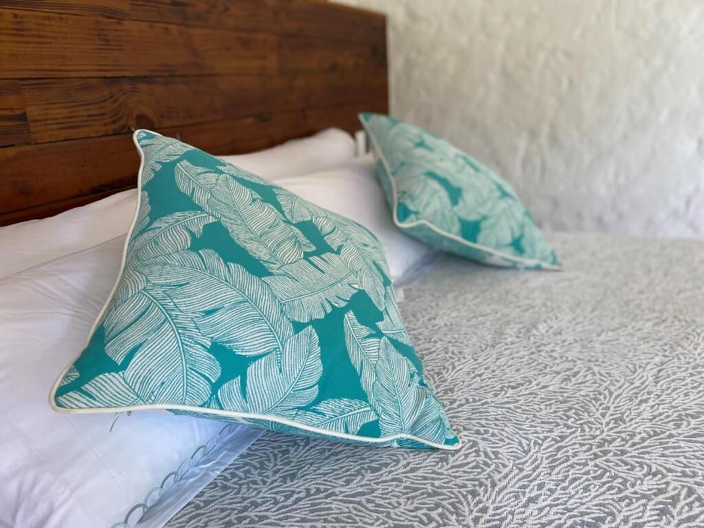 Cama com duas almofadas estampadas com padrão de folha, cabeceira de madeira e colcha cinza texturizada.