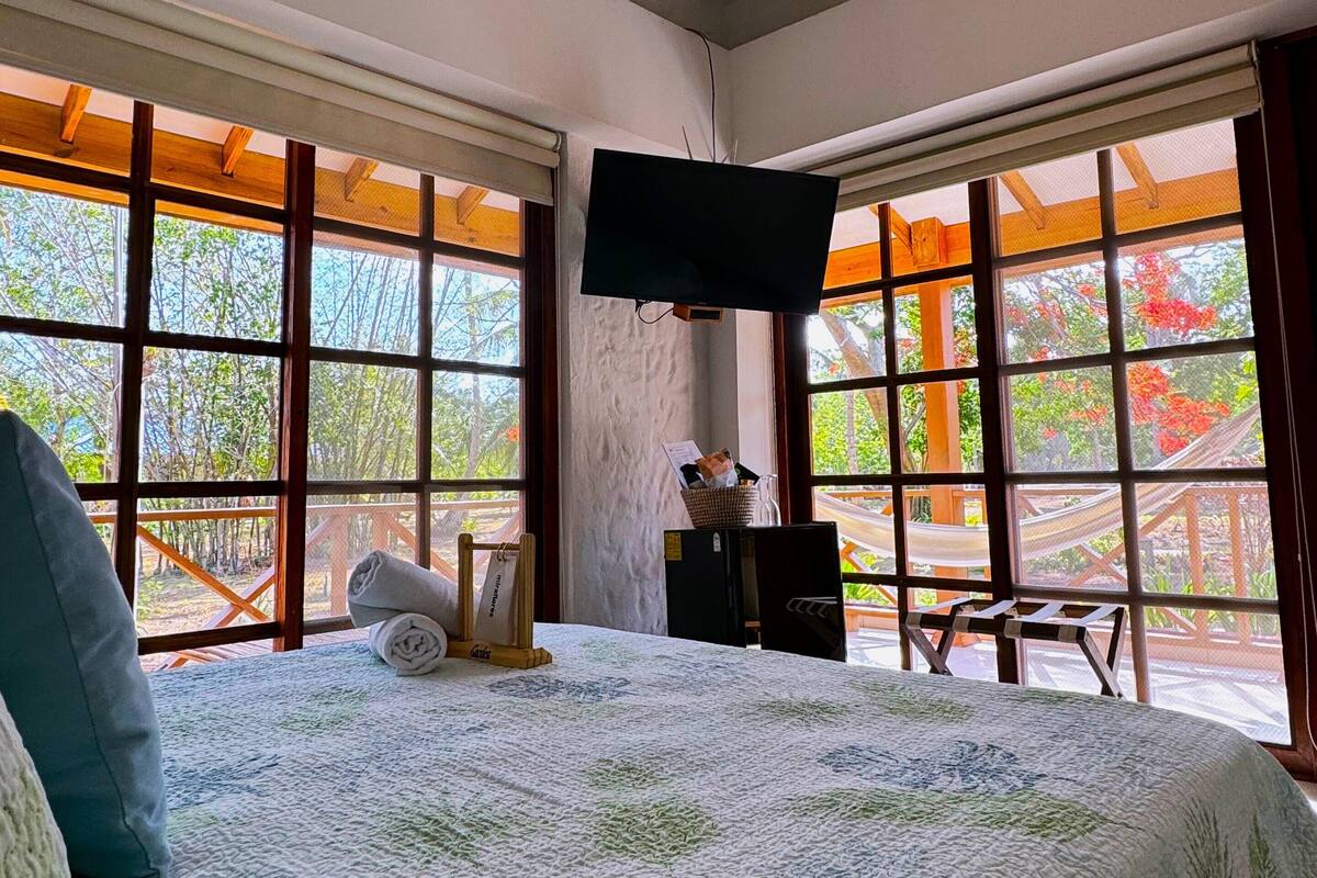 Un dormitorio con una cama grande, vigas de madera, un televisor de pantalla plana y ventanas amplias con vistas a árboles y una terraza de madera.