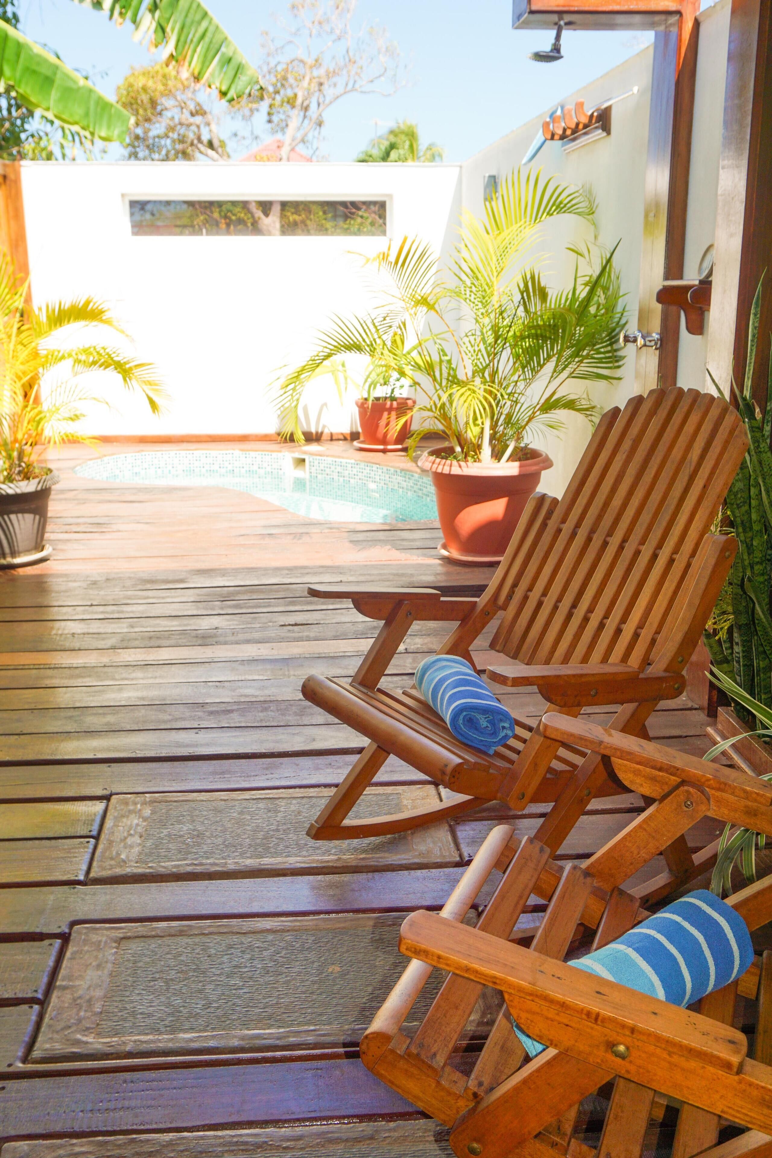 Una terraza de madera con una piscina, plantas en macetas y una silla de madera con cojín azul.