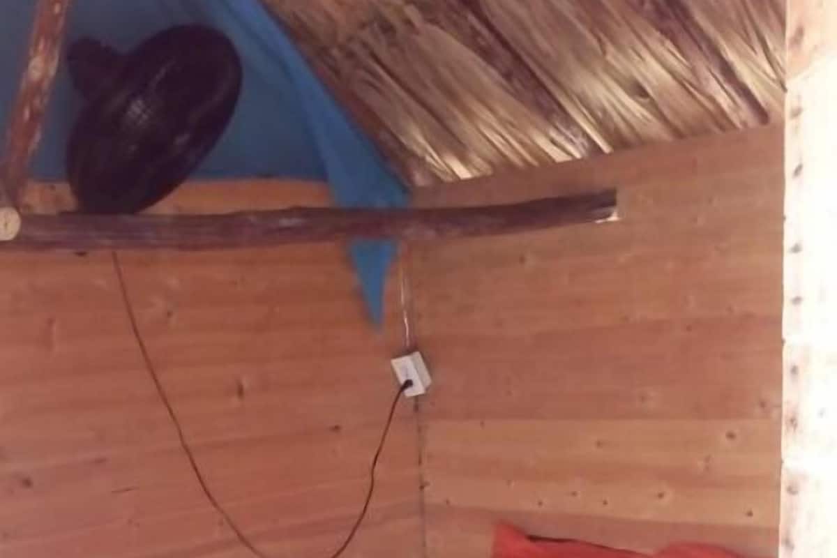 A small, single-bed cabin with a wooden ceiling and walls, a simple bed with an orange mattress, and a power outlet on the wall.