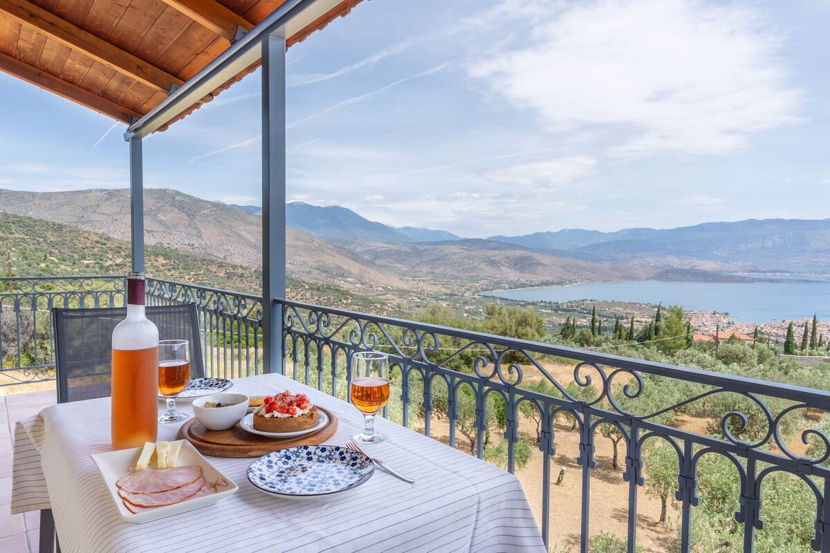 A table set with food and drinks on a balcony overlooking a mountainous landscape and a body of water.