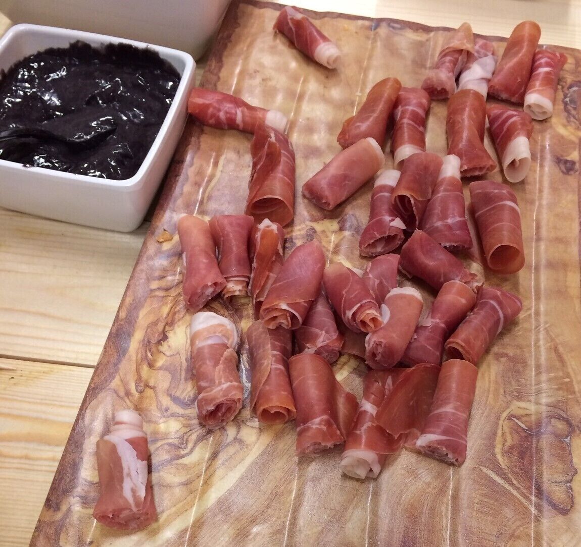 Une planche à découper en bois sur laquelle sont disposées des tranches de charcuterie et un bol contenant une sauce noire.