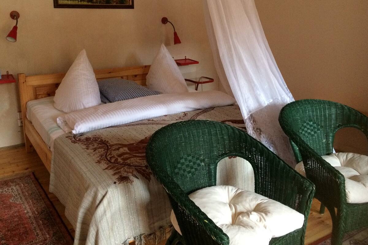 A bedroom with a bed, pillows, a canopy, two green wicker chairs, a red lamp, and a painting on the wall.