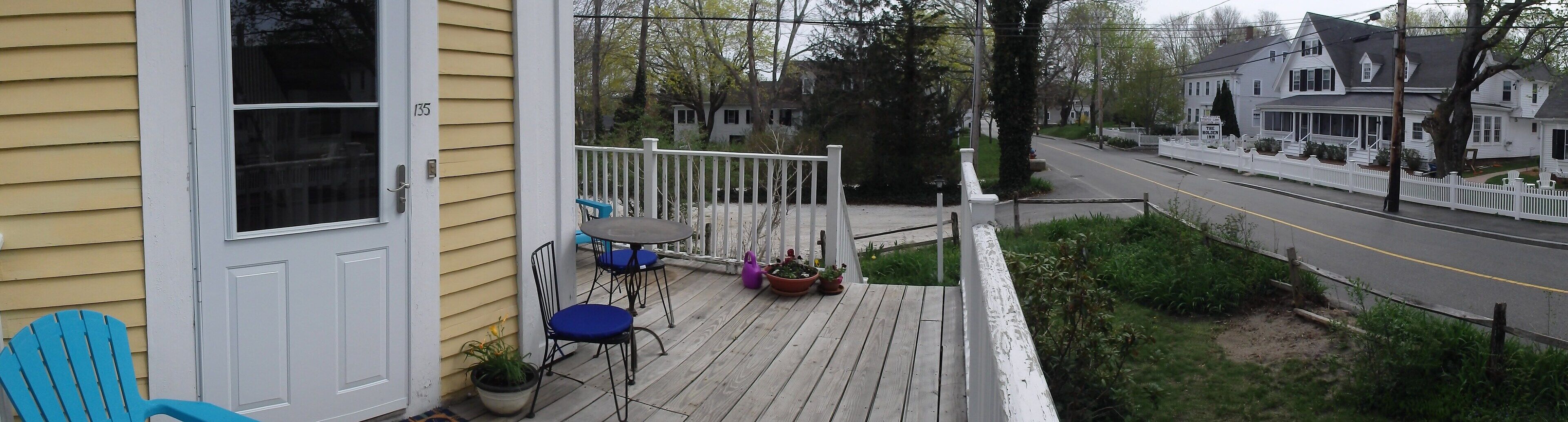 A wooden deck with outdoor seating, a white picket fence, and a road in the background.