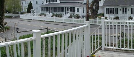 A white picket fence, a wooden deck, and a road in front of a building with a porch.