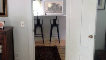 Un dormitorio con cama, suelo de madera, puerta blanca y un pasillo que da a una zona de cocina con dos taburetes y un cuadro de paisaje urbano.