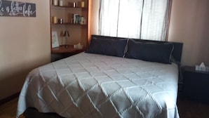 Ein Schlafzimmer mit einem Bett, einem Nachttisch, einem Regal mit Büchern und Kerzen sowie einem Fenster mit Vorhängen.