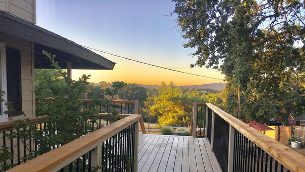 Una terraza de madera con barandilla metálica, que da a una vista de un paisaje con árboles y una cadena montañosa distante.