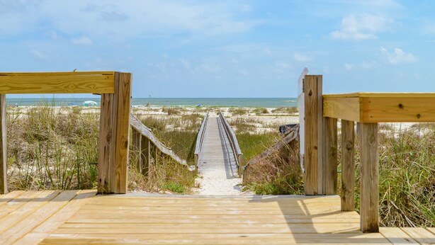 Ein hölzerner Steg führt zu einem Strand mit Dünen und blauem Himmel.