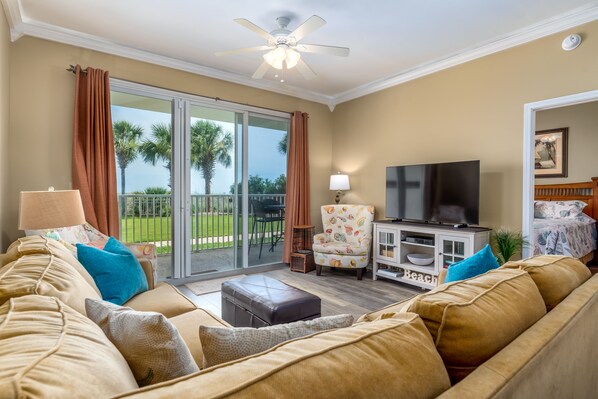 A living room with a sofa, a television, and a sliding glass door leading to a balcony with a view of palm trees. - Footprints in the Sand - 1st Floor Walk-out unit at the Inn on Dauphin Island (Dauphin Island)