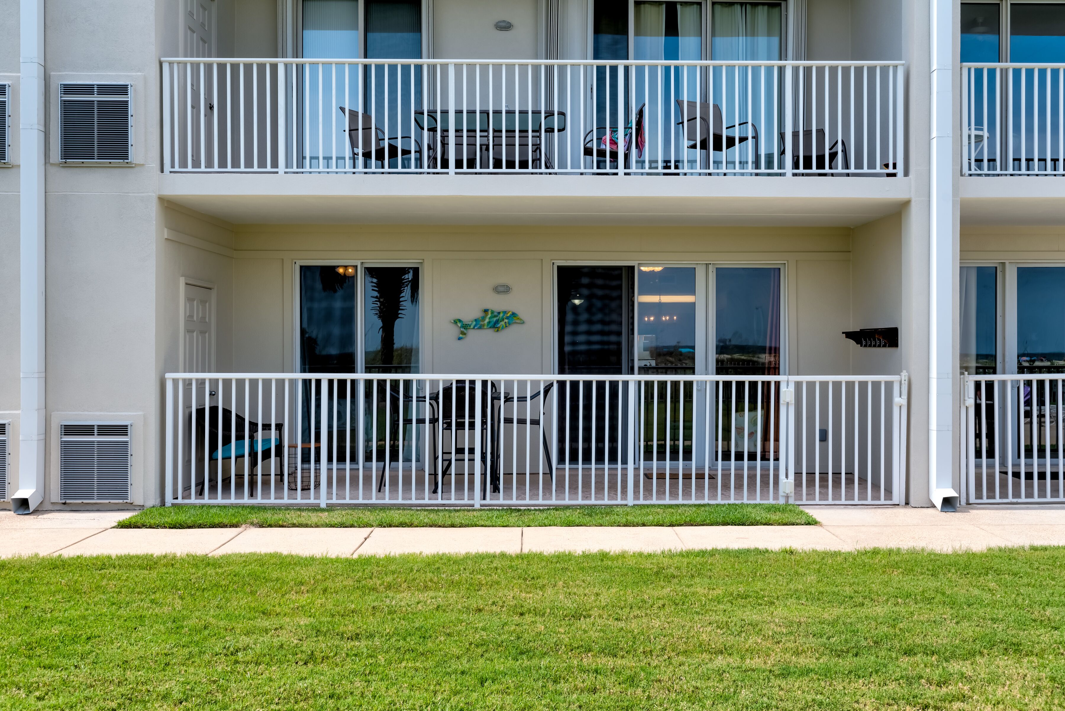 A multi-story building with balconies, large windows, and a grassy area in front.