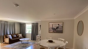 A spacious living room with a wooden floor, a brown leather sofa, a white dining table with chairs, and a painting on the wall.