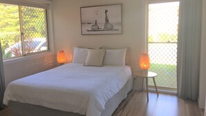 A bedroom with a bed, two bedside lamps, a ceiling fan, a window with a view, and a painting on the wall.