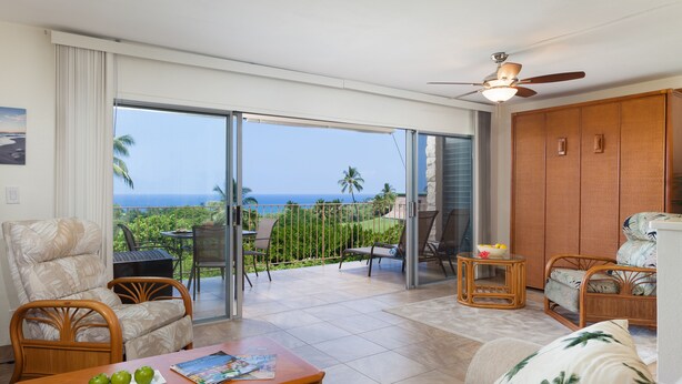 A spacious living area with a view of the ocean, a ceiling fan, and a dining table with apples. - Sunset Hale Kona: Ocean, Golf Course View, Keauhou Bay, Kona, Sleeps 4 (Kailua-Kona)